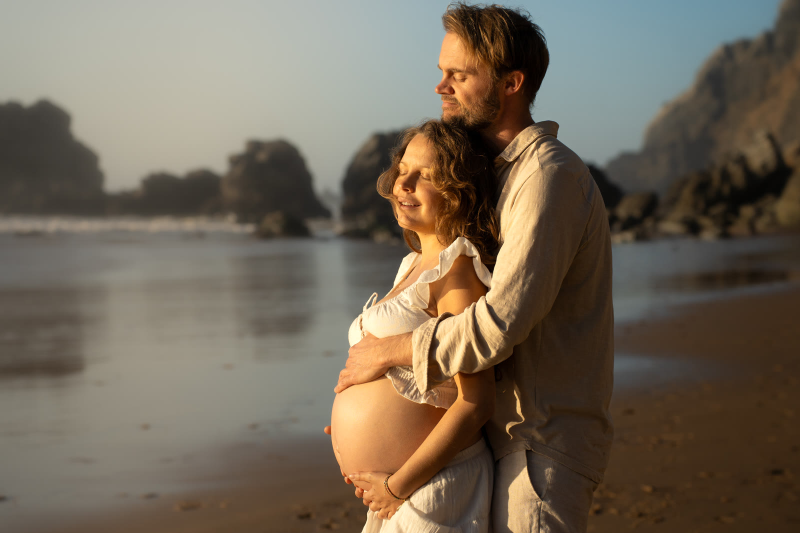 Pregnant woman held by her partner on the beach at golden hour